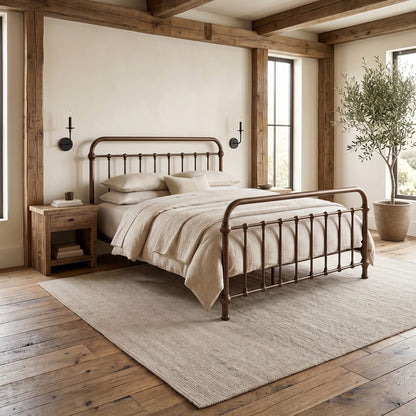Heiressy 20th c Americana Iron Bed in Copper Wash finish with a curved headboard and footboard, styled in a warm, neutral-toned bedroom with a matching nightstand, textured rug, and wooden ceiling beams.