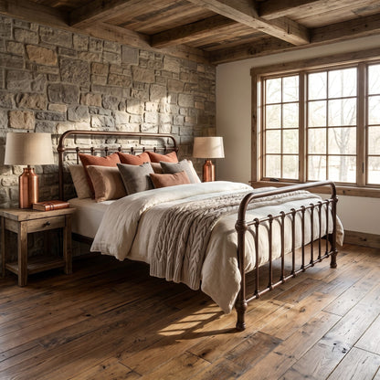 Heiressy 20th c Americana Iron Bed in Restored Copper. A rustic bedroom scene featuring a metal bed frame with beige bedding, styled with earthy accent pillows, set against a stone wall and a large multi-pane window.