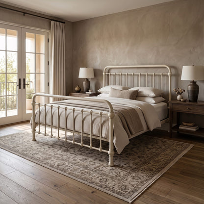 Heiressy 20th c Americana Iron Bedf in Restored Ivory Finish. A classic white metal bed frame with a tall headboard and footboard stands on a patterned area rug in a beautifully styled bedroom with beige walls, wooden floors, and matching nightstands.