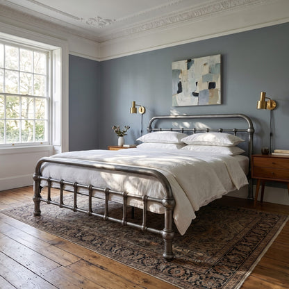 20th c Americana Low Profile Iron Platform Bed by Heiressy.  A metal bed frame with a curved footboard and headboard stands in a styled bedroom with white bedding, a patterned area rug, wooden floors, and blue-gray walls.