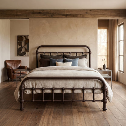 20th c Americana Low Profile Iron Platform Bed by Heiressy. A dark metal bed frame with a curved headboard and footboard, styled with neutral linens and blue accent pillows in a rustic, warm-toned bedroom.