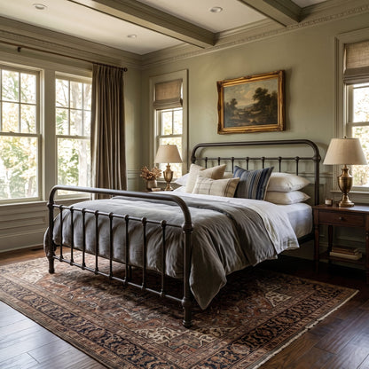 Heiressy 20th c Americana Iron Bed in Stond Ground Finish. A classic bedroom scene featuring a black metal bed frame with gray bedding, styled on a patterned area rug. The room has sage green walls, wooden ceiling beams, and large windows with brown drapes.