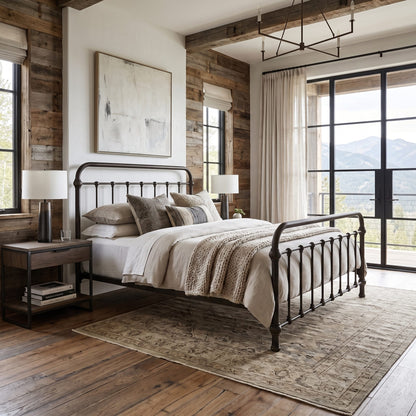 Heiressy 20th c Americana Iron Bed in Tennery Black Finish. A rustic bedroom featuring a black metal bed frame with a curved footboard, styled with neutral-toned bedding, a patterned area rug, and a dark wood nightstand. The room has wood-paneled walls, a large window with a view of mountains, and a modern geometric chandelier.