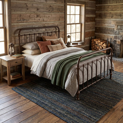 Heiressy 20th c Americana Iron Bed with a Timeworn Rust headboard and footboard, styled in a cabin bedroom with wood-paneled walls, a nightstand, windows, and a large blue-gray woven rug.