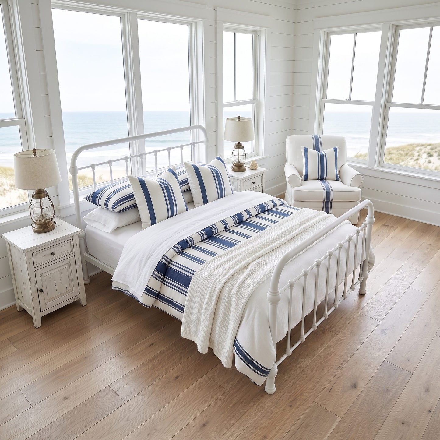 A bright coastal bedroom with an Heiressy 20th c Americana Iron Bed in White Wash. The bedroom has a white metal bed frame, white bedding, and blue-and-white striped pillows and throw, set in a sunlit room with wood floors, matching armchair, and large windows overlooking the ocean.